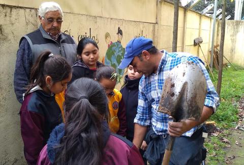Un grupo de niños escucha atentamente a un adulto que les enseña a usar una pala en un entorno al aire libre.