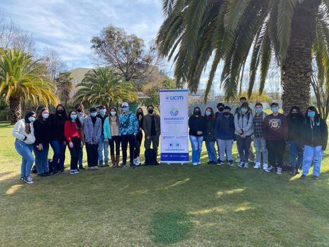 Un grupo de estudiantes posando juntos en un área verde con palmeras y un cartel de la UCM en el fondo.