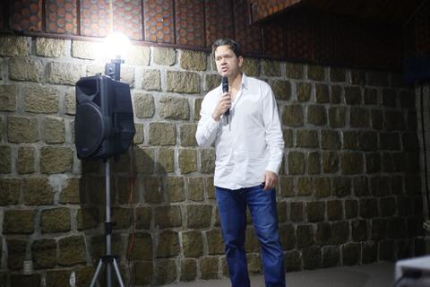 Un hombre de pie frente a un micrófono en un escenario con pared de piedra al fondo.