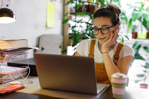 Una mujer sentada frente a una computadora portátil en un ambiente con plantas.