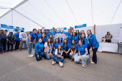 Un grupo de jóvenes posando en un evento bajo una carpa, vistiendo chaquetas azules y sonriendo.