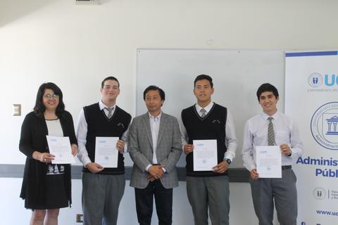 Un grupo de estudiantes posando con diplomas junto a un profesor en un aula.