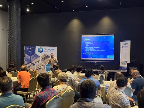 Una conferencia con asistentes sentados frente a un ponente que presenta en una pantalla.