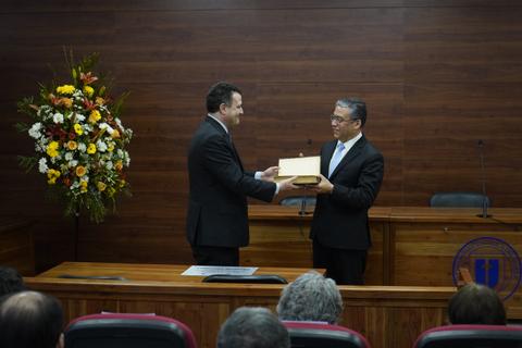 Dos hombres en un evento formal intercambian un objeto en un ambiente de oficina decorado con flores.