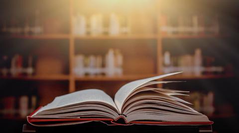 Un libro abierto descansa sobre una mesa, con un fondo de estanterías difusas.