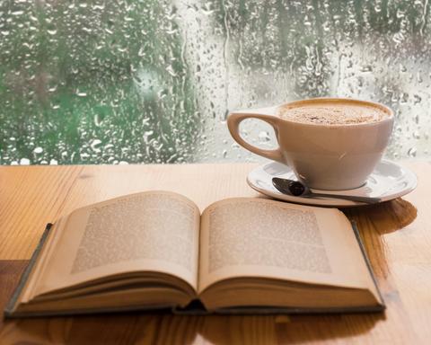 Una taza de café humeante junto a un libro abierto sobre una mesa de madera, con la lluvia cayendo en la ventana de fondo.
