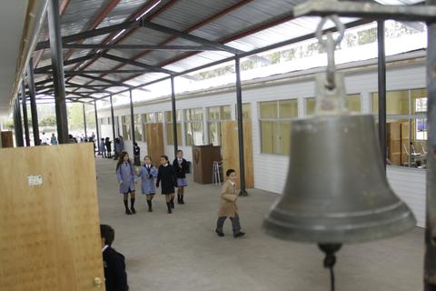 Un grupo de estudiantes camina por un pasillo escolar mientras una campana cuelga en primer plano.