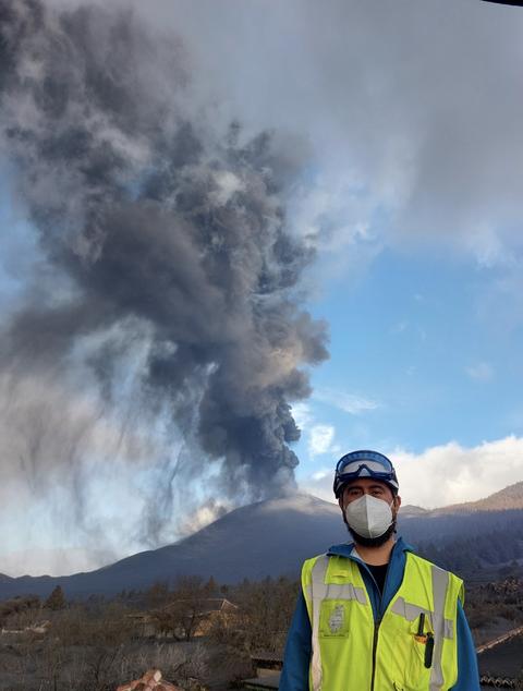 Un hombre con máscara facial y chaleco amarillo observa una erupción volcánica en el fondo.