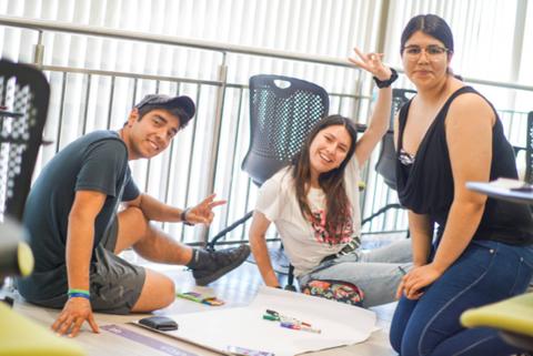 Un grupo de tres personas se divierte mientras están sentados en el suelo, rodeados de papeles y utensilios de dibujo.