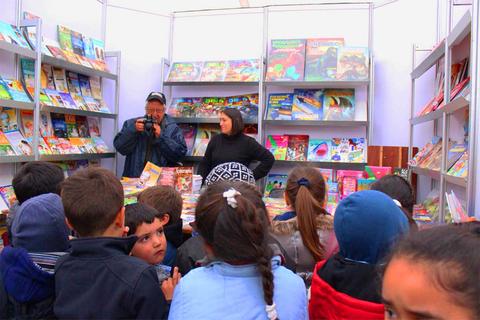 Un grupo de niños está observando una presentación en un evento literario, mientras un hombre toma fotos y una mujer los acompaña.