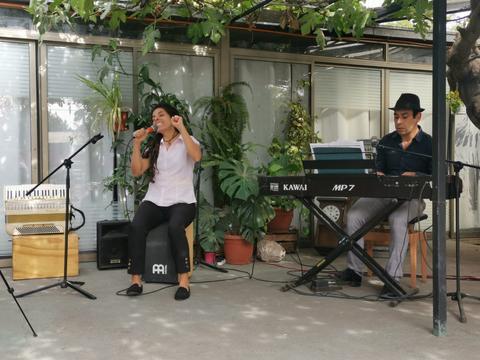 Una cantante interpreta una canción mientras un pianista la acompaña en un ambiente lleno de plantas.
