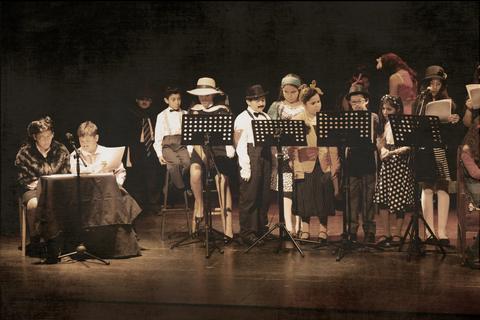 Un grupo de niños y adultos se presenta en un escenario durante una actuación teatral con vestimenta variada.