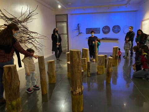 Un grupo de personas, incluyendo niños, explora una instalación artística con troncos en un ambiente iluminado con luz azul.
