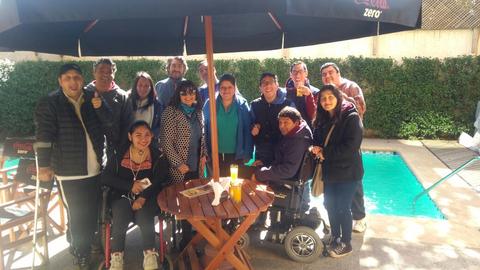 Un grupo de personas sonrientes se reúne alrededor de una mesa al aire libre cerca de una piscina.