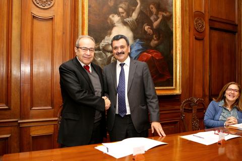 Dos hombres se dan la mano en un entorno formal, con una pintura en la pared detrás de ellos.