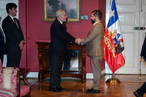 Dos personas se están dando la mano en una ceremonia oficial con una bandera chilena de fondo.