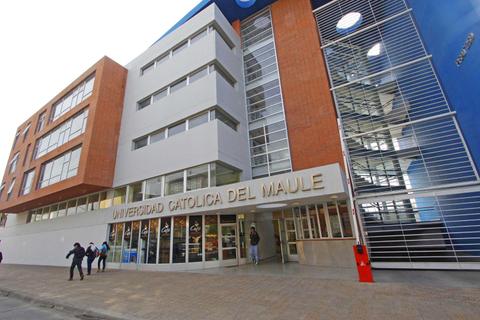 La imagen muestra la fachada de la Universidad Católica del Maule con un diseño moderno y varias personas caminando frente a ella.
