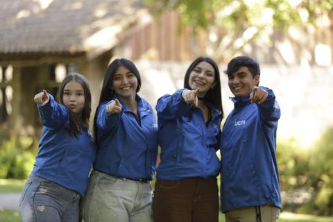 Un grupo de cuatro jóvenes sonríe y señala hacia la cámara en un entorno al aire libre.