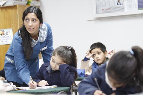 Una maestra se muestra atenta mientras los alumnos trabajan en sus escritorios.