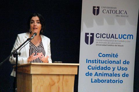 Una mujer presenta en un evento en la Universidad Católica del Maule, junto a una pancarta del Comité Institucional de Cuidado y Uso de Animales de Laboratorio.