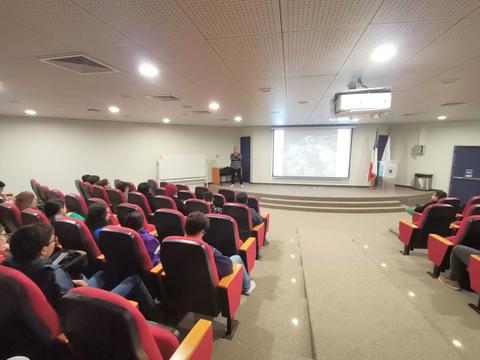 Un aula con asientos rojos donde un presentador habla frente a una audiencia sentada.