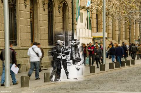 La imagen muestra una representación artística donde dos soldados en blanco y negro están contrastados con una multitud que camina en color en una calle moderna.