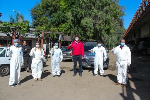 Un grupo de personas vestidas con trajes de bioseguridad se encuentra en un lugar al aire libre, mostrando un gesto de saludo.