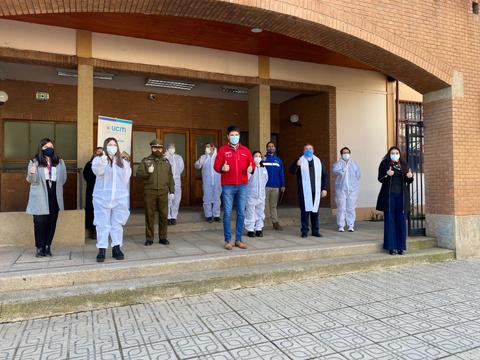 Un grupo de personas vestidas con trajes de protección se encuentra frente a un edificio, haciendo gestos de saludo y unidad.