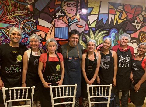 Un grupo de personas posan sonrientes en un ambiente colorido y creativo, con delantales de cocina y cofia en la cabeza.