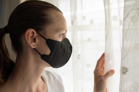 Una mujer con mascarilla observa por la ventana mientras toca las cortinas.