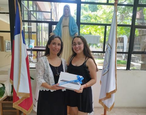 Una mujer y una joven posan juntas sosteniendo un regalo en un ambiente institucional con estatuas y banderas de fondo.