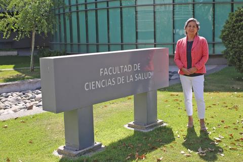 Una mujer sonriente se encuentra de pie junto a un letrero que indica la Facultad de Ciencias de la Salud.