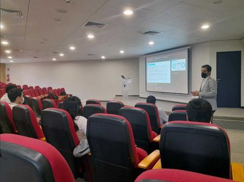 Un presentador expone frente a un grupo de personas en un auditorio con asientos rojos.