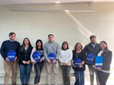 Un grupo de ocho personas posando en un entorno interior, todos sosteniendo mochilas azules con el logo de UCM.
