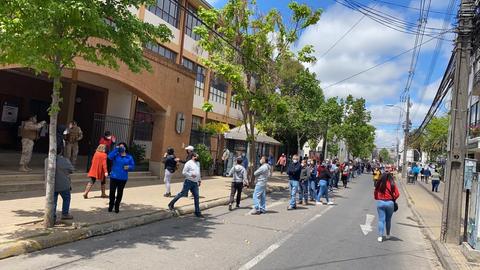 Una multitud de personas espera en una fila afuera de un edificio en un día soleado.