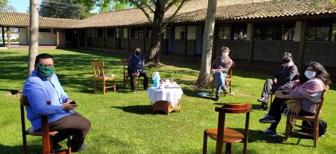 Un grupo de personas sentadas en sillas alrededor de una mesa en un ambiente al aire libre, rodeadas de árboles y edificios.