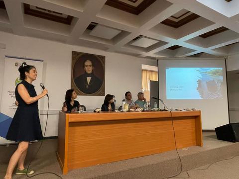 Una presentación en una sala con un ponente de pie y varios participantes sentados junto a una mesa.