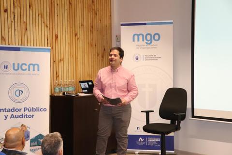 Un conferencista presenta un tema en un auditorio con banderas y logos de instituciones educativas.