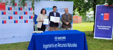Un grupo de tres personas sostiene un documento frente a una mesa con un mantel que lleva el logo de la UCM en un entorno al aire libre.