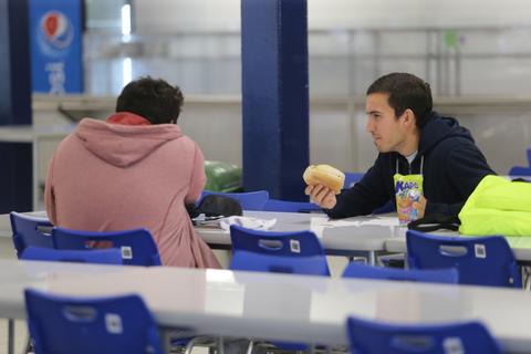 Dos jóvenes sentados en una mesa de cafeteria, uno de ellos come un bollo mientras el otro está de espaldas.
