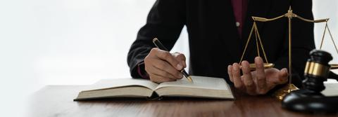 Un abogado escribiendo en un libro mientras sostiene una balanza de la justicia.