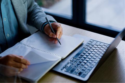 Una persona está escribiendo notas en un cuaderno mientras trabaja en su computador.