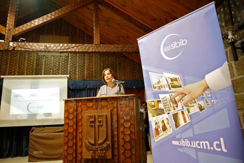 Una mujer se encuentra en un podio hablando frente a una audiencia en una sala decorada con madera.