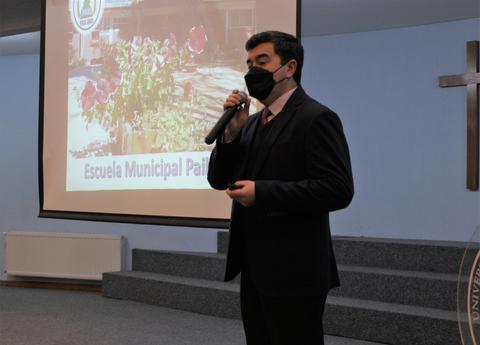Un hombre habla en un evento mientras lleva una mascarilla y hay una presentación detrás de él.