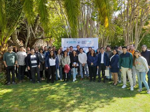 Un grupo de personas se reúne en un evento al aire libre, rodeado de árboles y un banner de la universidad.