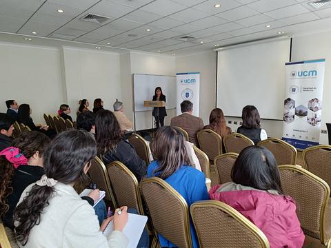 Una mujer presenta en un evento frente a una audiencia en un salón con sillas dispuestas para los asistentes.