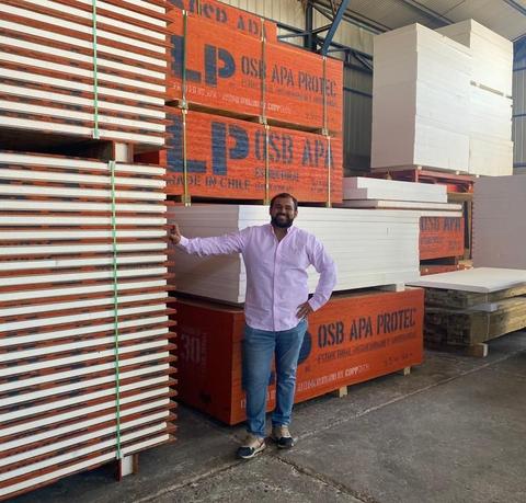 Una persona sonriente se encuentra junto a pilas de tableros de madera en un almacén.
