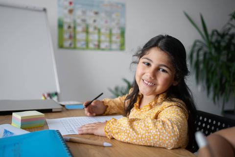 Una niña sonriente está sentada en una mesa, escribiendo en un cuaderno.
