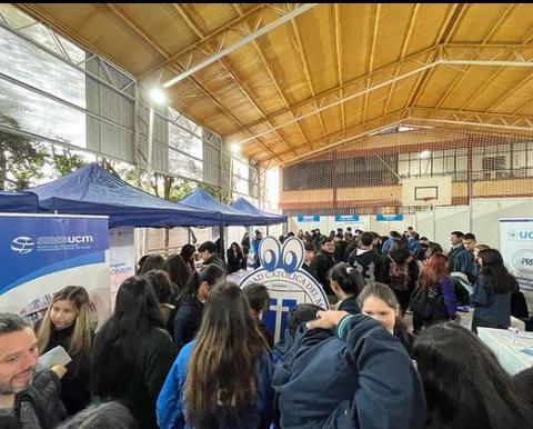 Una gran cantidad de estudiantes se reúnen en un evento dentro de un gimnasio, rodeados de stands y material informativo.