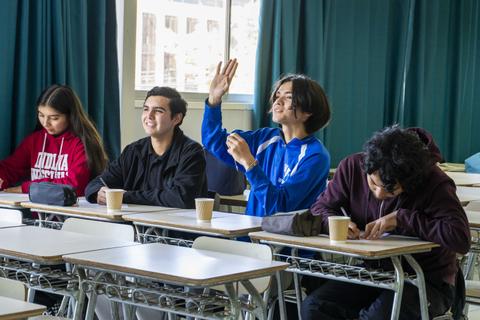 Un grupo de estudiantes en un aula, algunos levantando la mano y otros escribiendo.